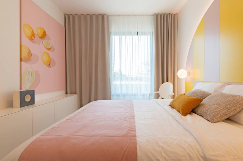 Spavaća soba sa bračnim krevetom, roza prekrivačem, bež zavesama i zidnom dekoracijom u pastelnoj paleti