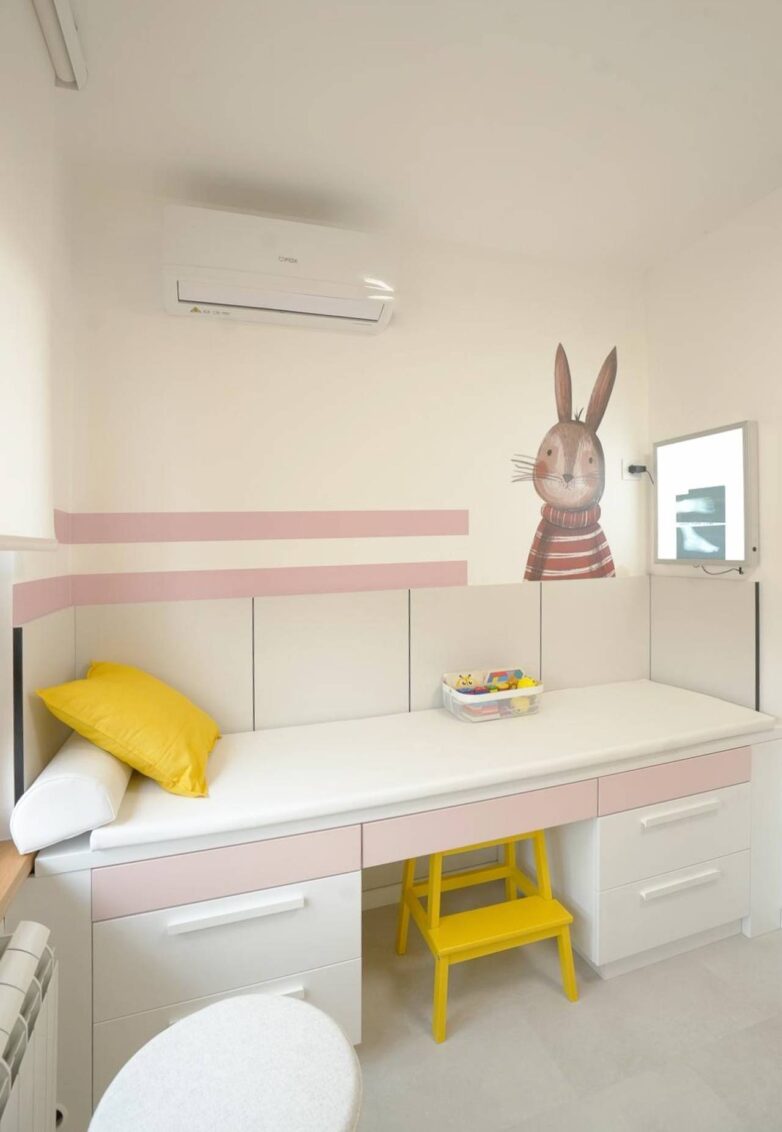 Pregledni sto sa fiokama u pedijatrijskoj ordinaciji, sa roze detaljima i ilustracijom zeke na zidu