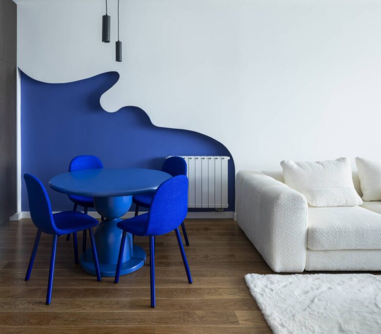 Trpezarija sa plavim okruglim stolom i stolicama ispred organski oblikovanog indigo zida u stanu u Banja Luci