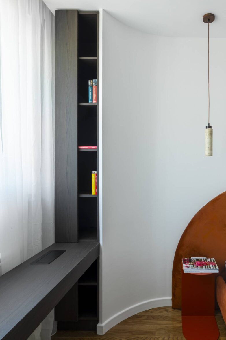 Radni kutak u stanu u Banja Luci sa tamnim radnim stolom, uskom bibliotekom i zakrivljenim zidom pored terakota uzglavlja