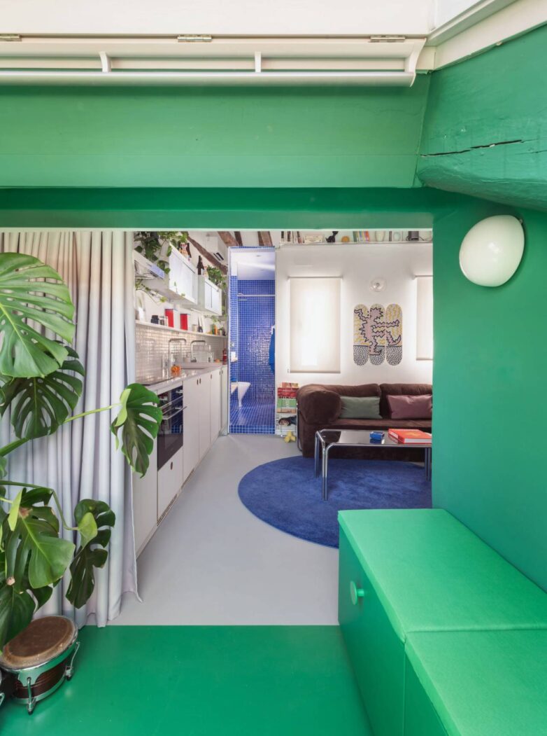 Pogled kroz zeleni deo potkrovlja ka dnevnoj sobi i kuhinji u stanu od 30 m2 u Madridu, sa plavim mozaik kupatilom u pozadini