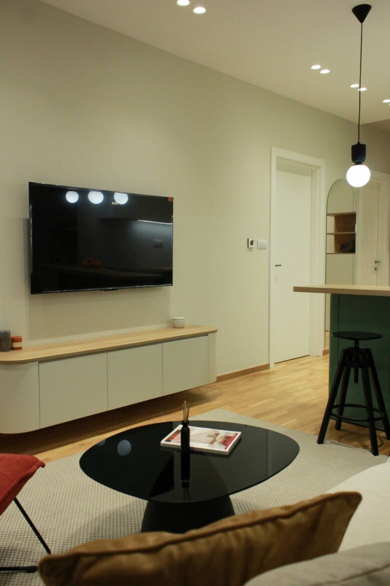 Dnevni boravak malog stana u Novom Sadu sa zidnim TV-om, niskim komodom i centralnim šankom