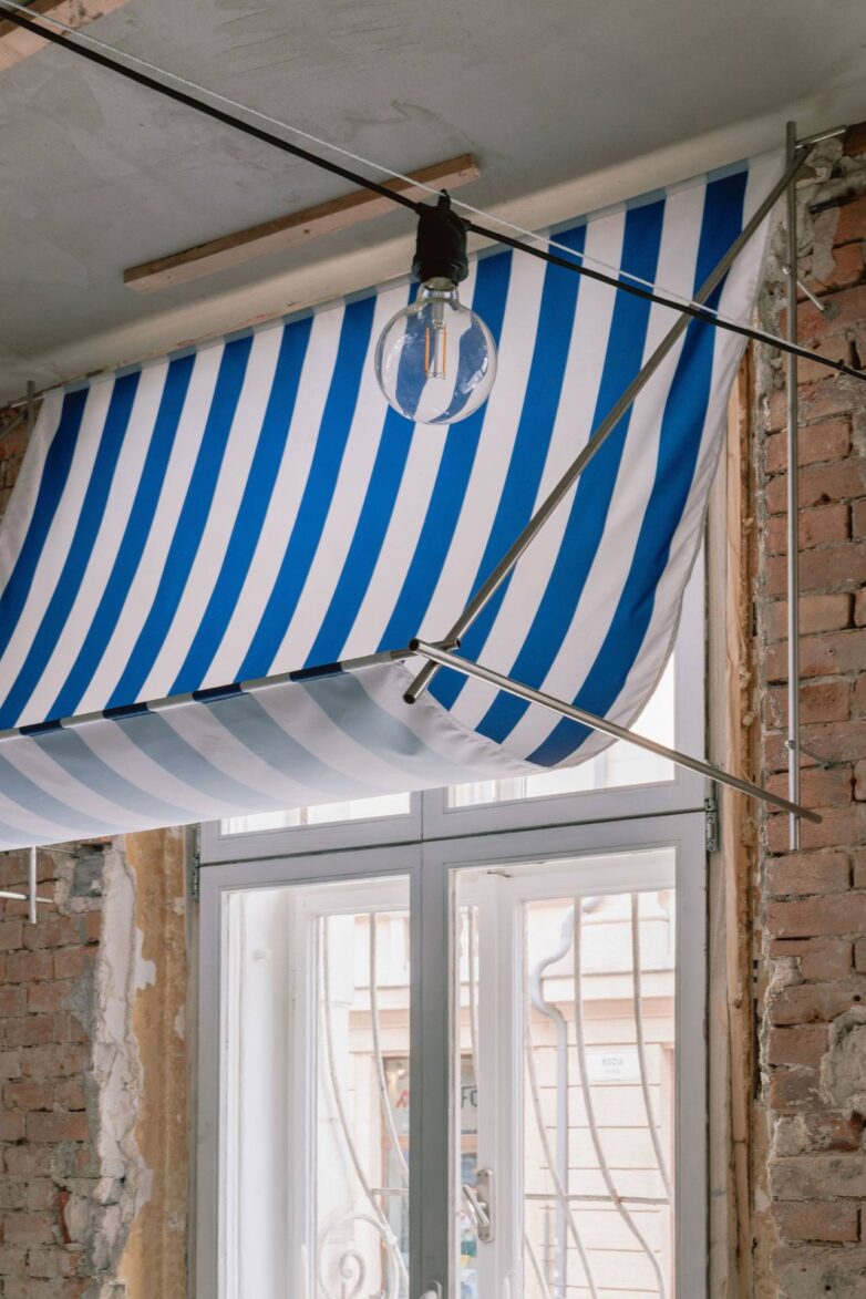 Plavo-bela prugasta tenda postavljena unutar prozora u enterijeru picerije Oli's Italiano u Bratislavi, sa vidljivom ciglom i industrijskom rasvetom