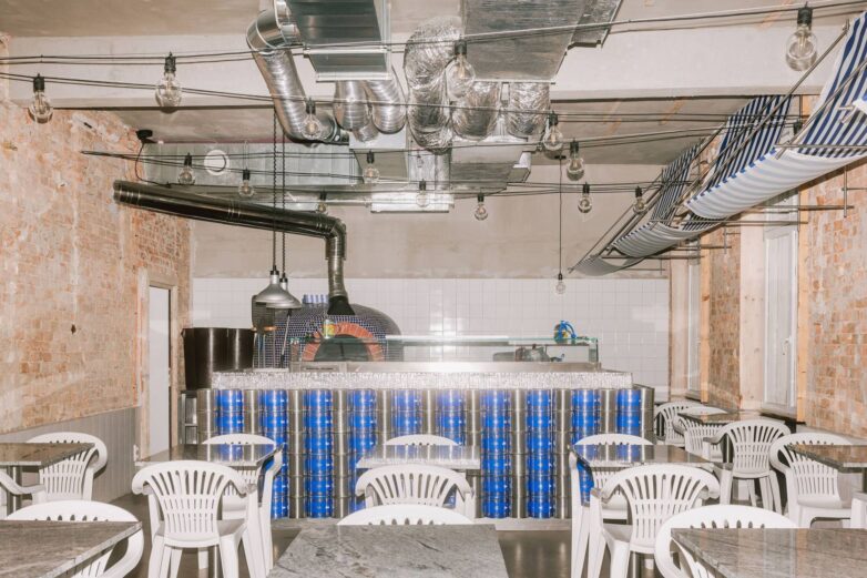 Peć za picu obložena plavim mozaikom u enterijeru picerije Oli's Italiano u Bratislavi sa belim Monobloc stolicama i industrijskim plafonom