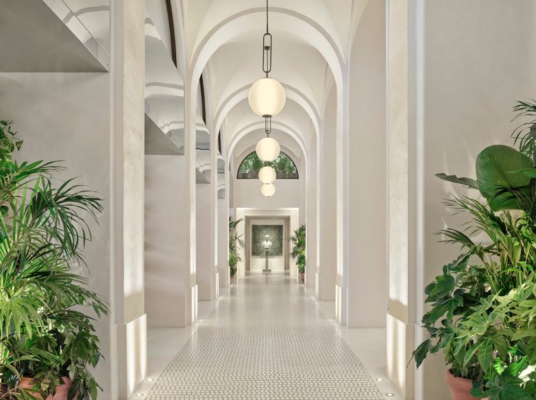 Dugačak hotelski hodnik sa lučnim svodovima, visećim svetlima i biljkama u saksijama, u svetlim neutralnim tonovima