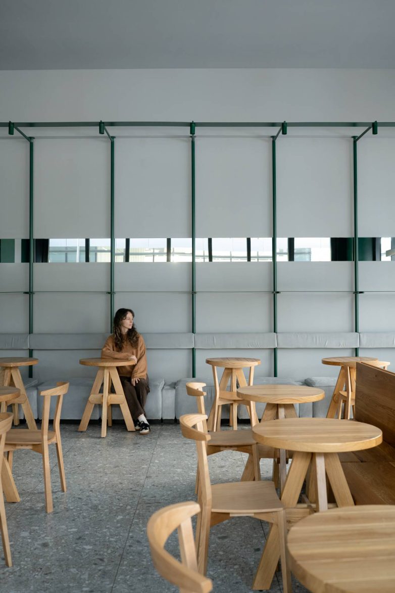 Zona sedenja u Kafeteriji u Čačku sa drvenim stolovima, tapaciranom klupom i zelenom metalnom konstrukcijom na zidu, projekat KIDZ Studio