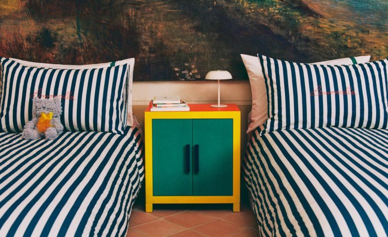 Dva kreveta sa prugastom posteljinom i natpisom Limoncella, razdvojena noćnim ormarićem u jarkim bojama