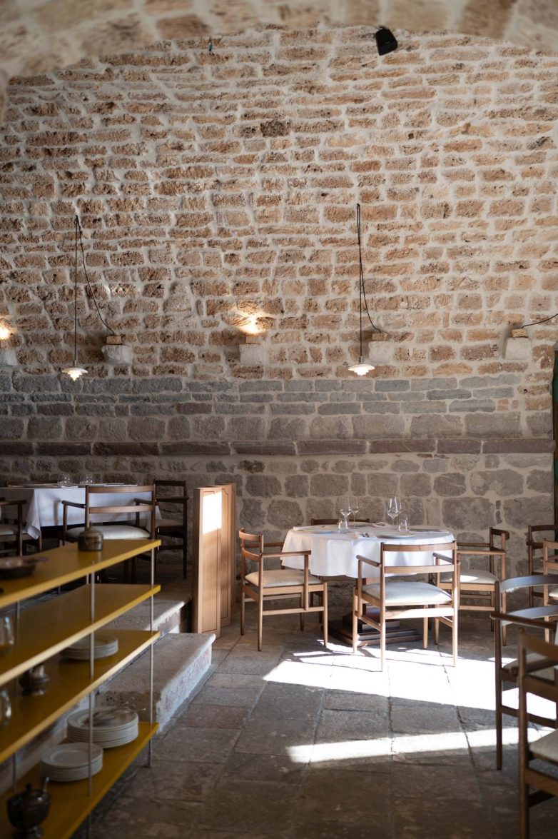 Restoranska sala sa kamenim zidom, drvenim stolicama i stolovima u restoranu Eho u Perastu