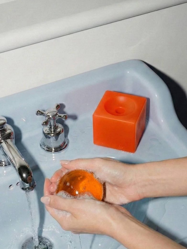 Narandžasti sferični sapun u rukama iznad lavaboa, sa kockastom bazom sa strane