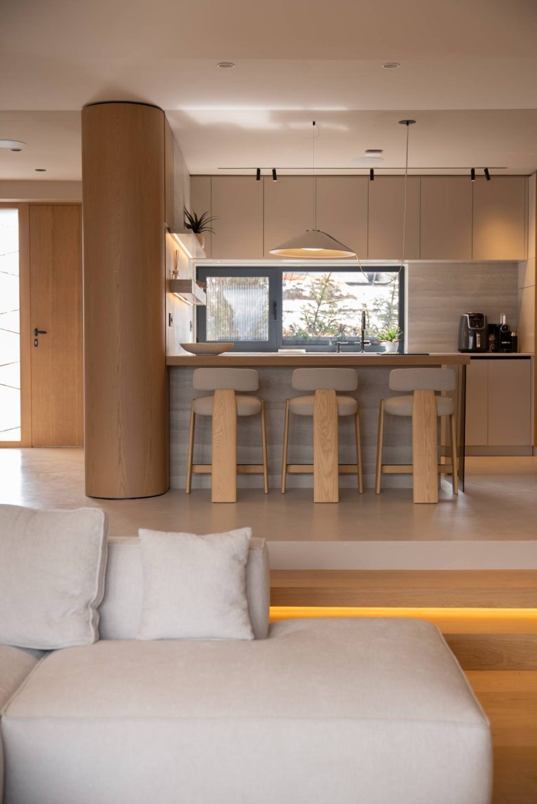 Kuhinja sa ostrvom, visokim barskim stolicama i kombinacijom drveta i neutralnih tonova u savremenom enterijeru