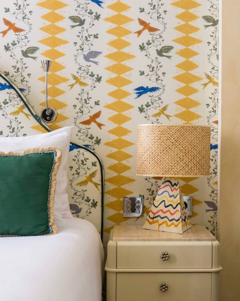 Noćni stočić sa lampom i uzglavljem kreveta ispred zida sa šarenim tapetama sa motivima ptica u hotelu Casa Monti u Rimu