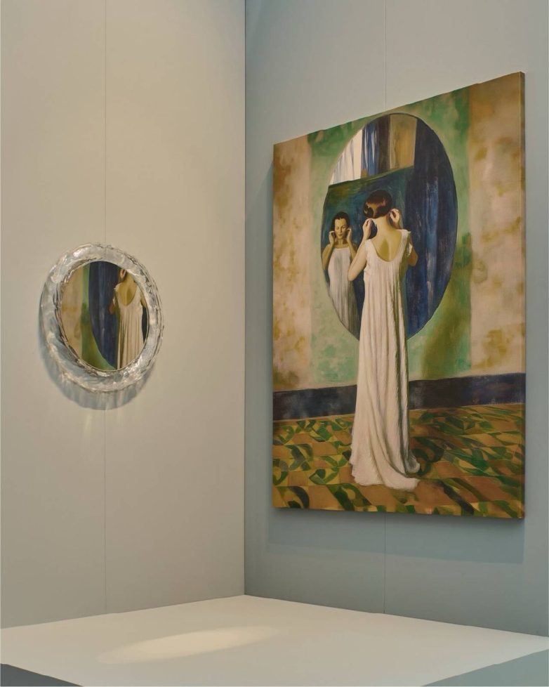 Svetli enterijer Kartell štanda sa velikom slikom žene ispred ogledala i dekorativnim ogledalom na zidu