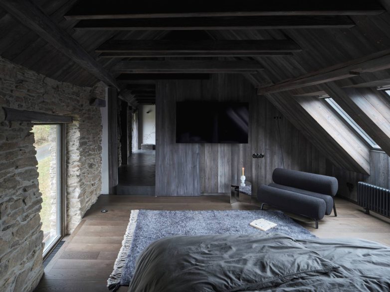 Spavaća zona u potkrovlju sa tamnim drvenim oblogama, kamenim zidom, krovnim prozorima i minimalističkim nameštajem