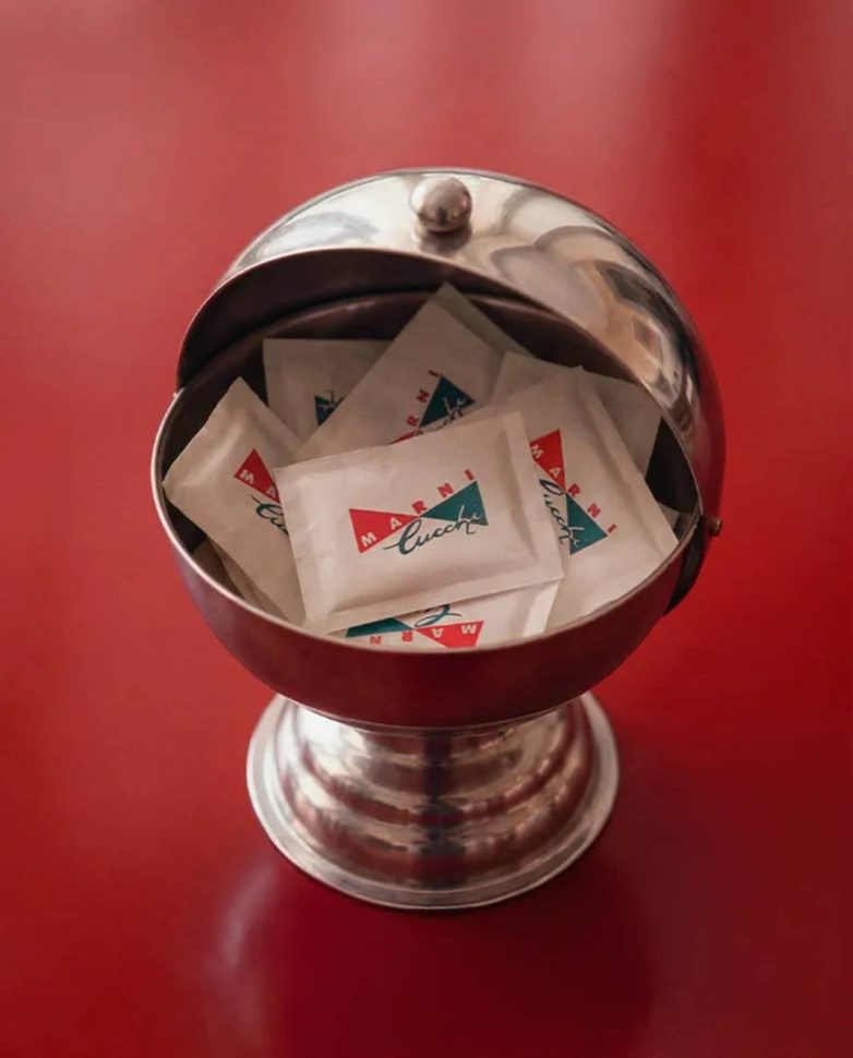 Metalna posuda sa kesicama šećera sa Marni i Cucchi logotipom na crvenoj podlozi