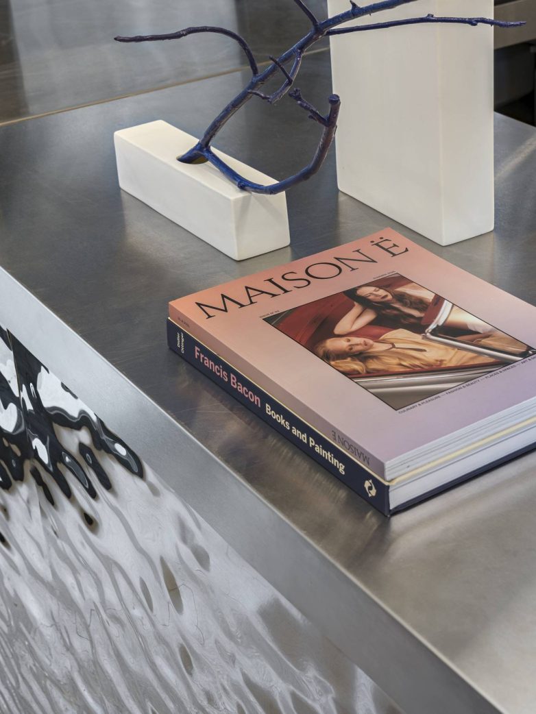 Metalni šank sa reflektujućom površinom na kojem se nalaze knjige i minimalistički dekorativni objekti sa plavom granom