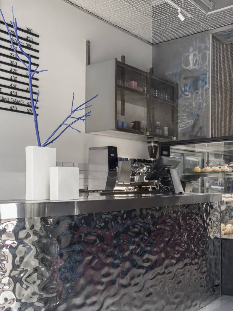 Barski deo kafića sa reflektujućim metalnim šankom, aparatom za kafu, vitrinom sa kolačima i minimalističkim policama na zidu