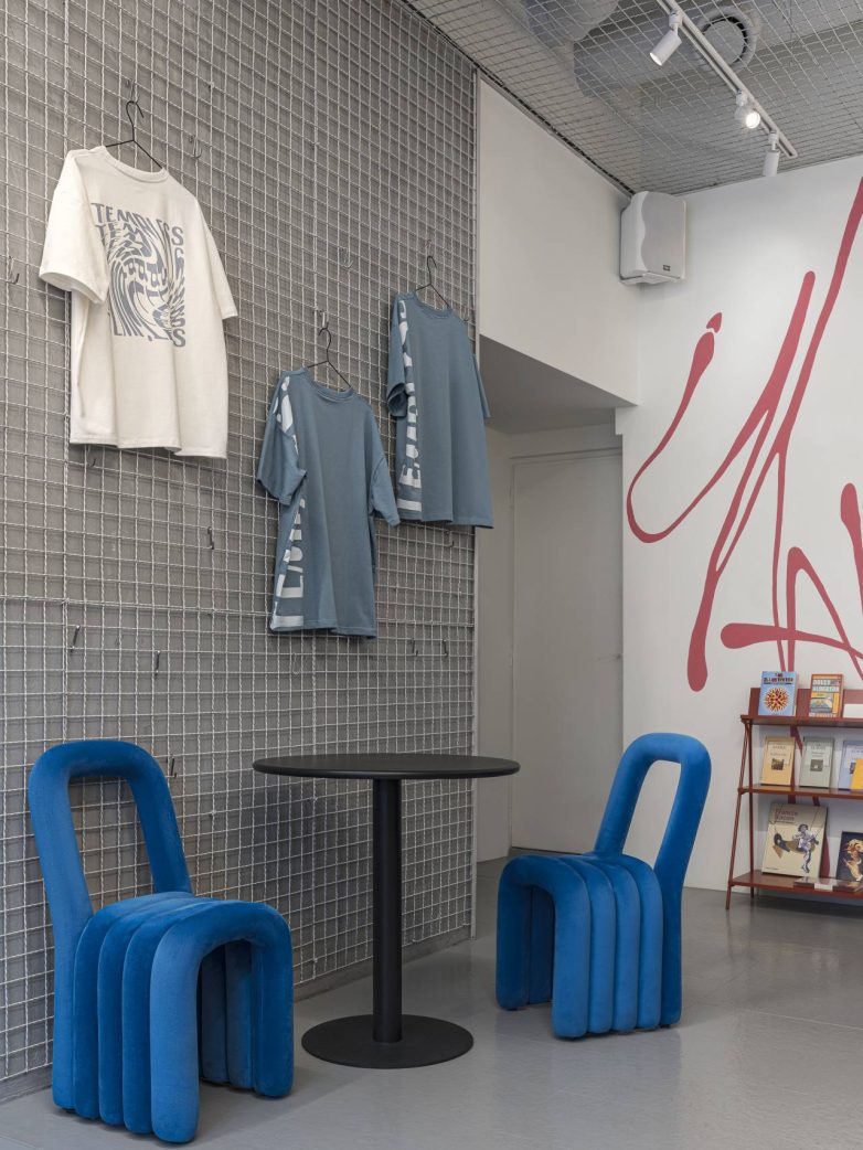 Detalj enterijera kafića sa zidom od metalne mreže na kojem vise majice, crnim okruglim stolom i dve plave skulpturalne stolice