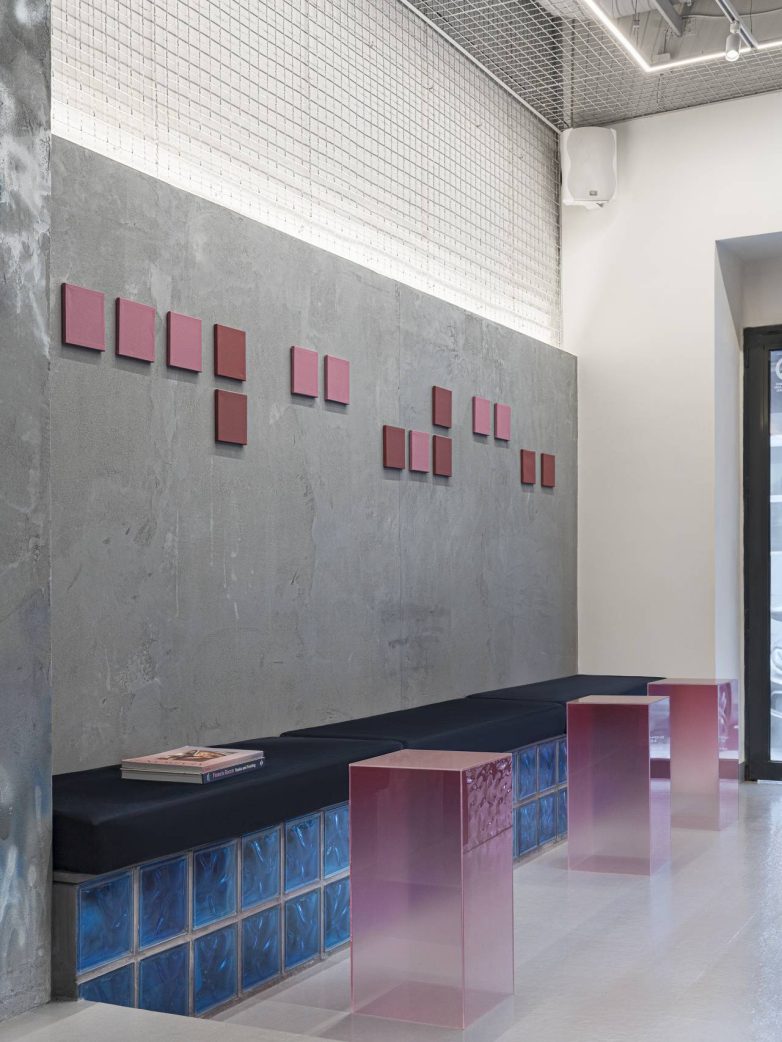 Minimalistički enterijer kafića sa dugom tapaciranom klupom, bazom od plavih staklenih blokova, roze poluprozirnim kubusima i sivim zidom sa geometrijskim umetničkim elementima