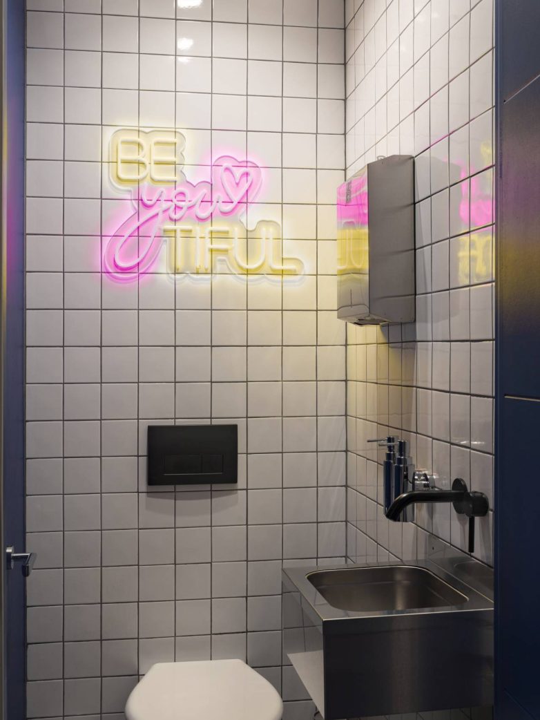 Mali toalet sa belim zidnim pločicama, neon natpisom u roze i žutoj boji, crnom slavinom i metalnim lavaboom