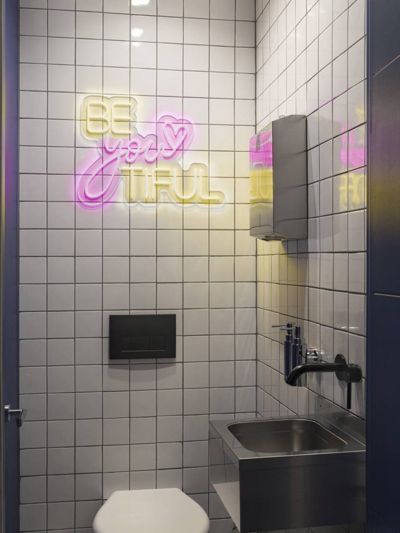 Mali toalet sa belim zidnim pločicama, neon natpisom u roze i žutoj boji, crnom slavinom i metalnim lavaboom