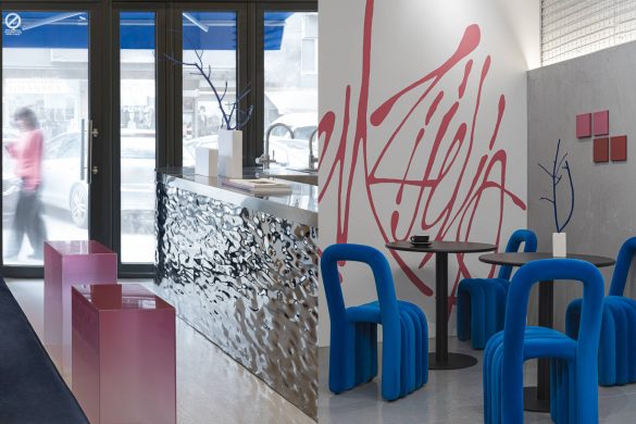 Ulazni deo malog kafića sa ogledalnim šankom koji reflektuje prostor, roze kubusima za sedenje i stolovima sa plavim skulpturalnim stolicama uz zid sa crvenim grafičkim natpisom