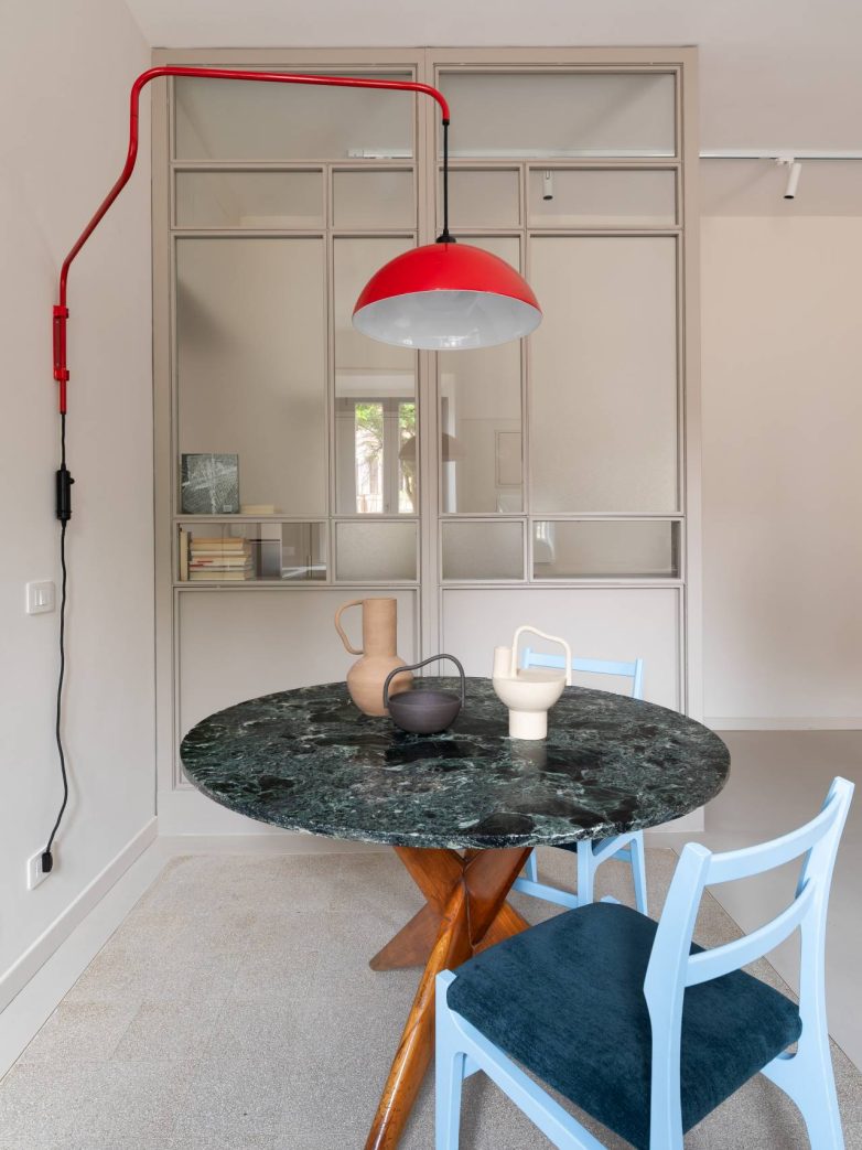 Trpezarijski deo stana Casa Garbatella u Rimu nalazi se ispred metalne staklene pregrade i ispod crvene zidne lampe.