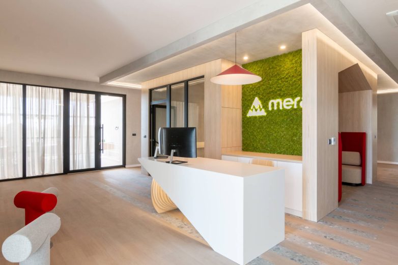 Prijemni pult u poslovnom prostoru kompanije Mera u Zrenjaninu nalazi se ispred zelenog zida sa logotipom, dok se u pozadini vidi "lounge" zona sa crvenim naslonima.