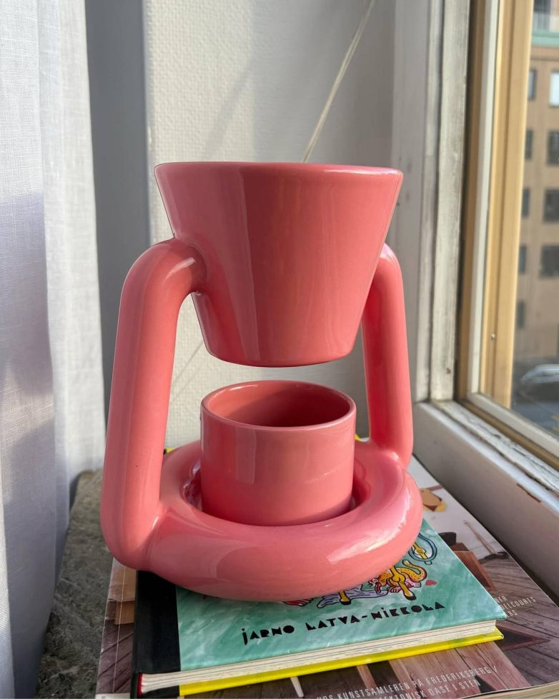 Roze Chunky Coffee Maker Gustafa Vestmana stoji pored prozora, postavljen na knjige i prikazan kao dekorativni predmet u enterijeru.