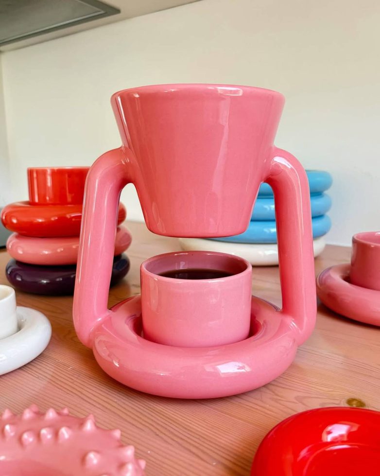 Roze keramički Chunky Coffee Maker Gustafa Vestmana prikazan je na drvenoj površini među šarenim predmetima iz iste kolekcije.