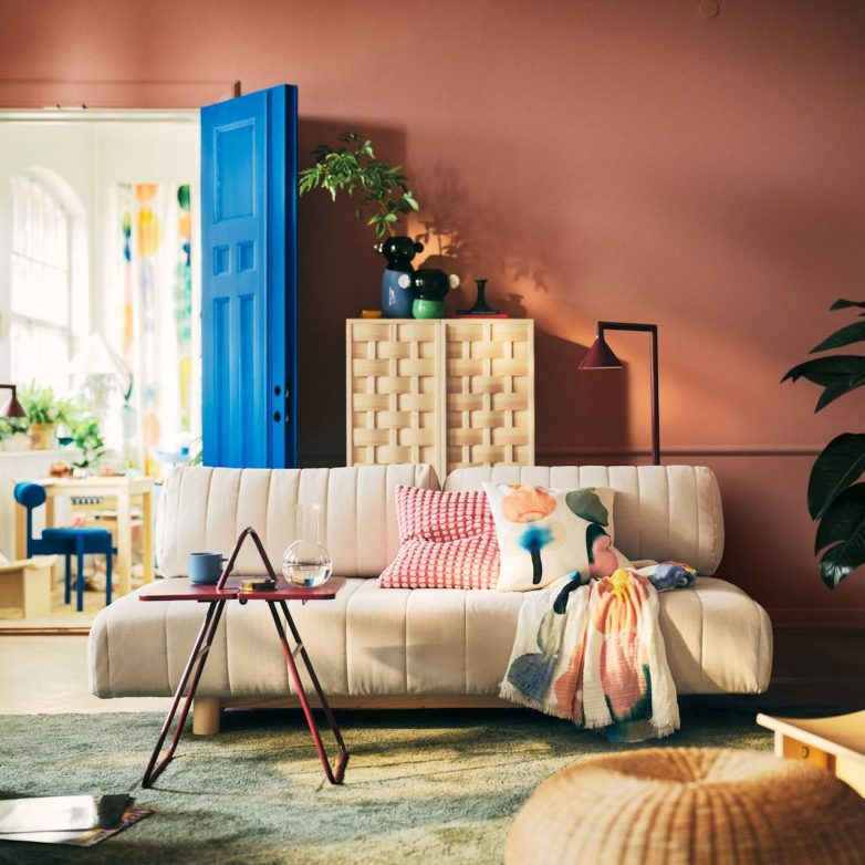 Svetla sofa iz IKEA PS 2026 kolekcije nalazi se u dnevnoj sobi sa plavim vratima, šarenim tekstilom i drvenim ormarićem