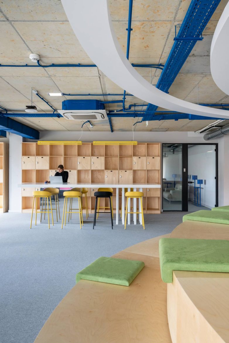 "Coworking" zona u kancelariji kompanije Serb Tech u Beogradu ima drvene ormariće, visoki sto sa žutim stolicama, zelene tapacirane elemente i otvorene instalacije na plafonu.