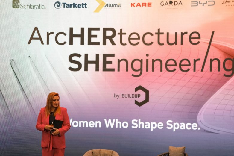 Voditeljka programa na konferenciji ArcHERtecture / SHEngineering stoji ispred velikog ekrana sa sloganom Women Who Shape Space.
