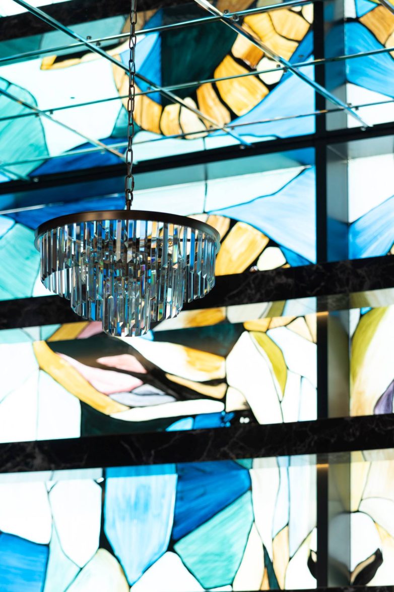 Detalj restorana GOAT u Banjoj Luci sa visećim lusterom i osvetljenom zidnom dekoracijom nalik vitražu u plavim, zelenim i žutim tonovima, projekat studija JB architect