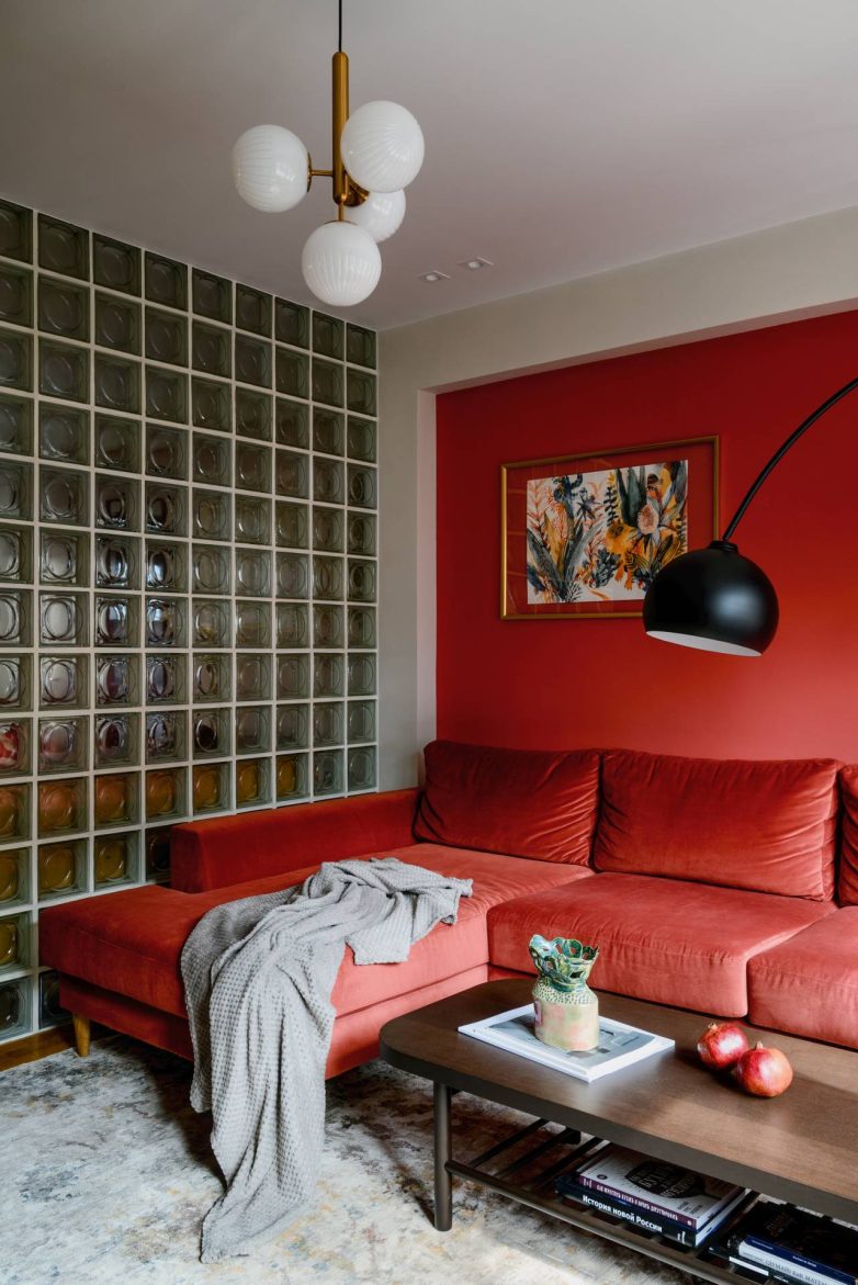 Crvena sofa u dnevnoj sobi sa zidom od staklenih blokova, podnom lampom i dekorativnim lusterom